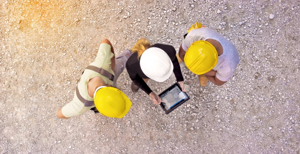 Female architect and two construction workers checking plans on digital tablet outside at construction site and pointing at progress on building.